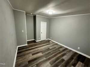 An empty room with gray walls and wood-look flooring. The room features a white door, a ceiling light fixture, and a small closet area to the left of the door.