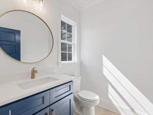 A modern bathroom with a round mirror above a blue vanity with a white countertop. A brass faucet and a white toilet are visible. Natural light streams through a window, casting shadows on the white walls and light wood floor.