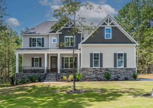 A large two-story house at 3407 River Manor Court, Franklinton, boasts a white and dark gray exterior with a gabled roof and stone accents. It features a covered front porch with columns, surrounded by lush greenery and trees under a clear blue sky.