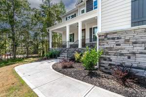 A modern house with a stone exterior and white siding. It features a front porch with black railings and steps leading to the entrance. The yard has a curved concrete path and landscaping with shrubs and plants. Trees are visible in the background.