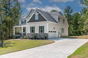 A two-story house with a gray roof, white siding, and a stone facade. It features a front porch, multiple windows, and a two-car garage. The house is surrounded by a lawn and trees under a partly cloudy sky.