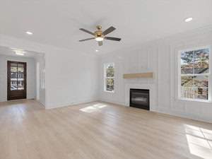 A bright, empty living room with light wood floors, white walls, and a ceiling fan. There are two large windows and a fireplace with a minimal wooden mantle. The room is well-lit by natural light.