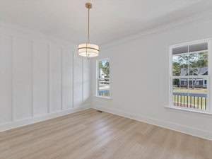 A bright, empty room with light wood flooring and white walls, featuring a modern circular pendant light. Two windows with views of a suburban street and houses allow natural light to fill the space.
