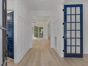 A hallway with light wood flooring, white walls, and a blue glass-paneled door on the right. It leads to a bright living space with large windows and a glimpse of an outdoor area. The space has a modern and minimalist design.
