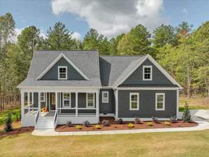 Located at 3405 River Manor Court in Franklinton, this charming two-story gray house with white trim is nestled among trees. It boasts a large front porch with chairs, a neatly landscaped yard, and enjoys the backdrop of a clear blue sky.