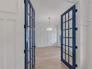 A room with light wooden floors and white walls is seen through open blue double glass doors. A modern chandelier hangs from the ceiling, and a window is on the right wall, allowing natural light into the space.