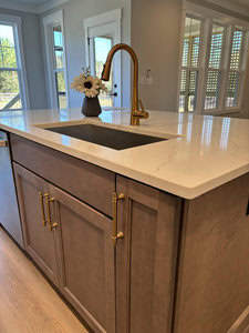A kitchen island with a marble countertop, featuring a gold faucet over a sink. The wooden cabinetry below has golden handles. A vase with white flowers sits on the countertop. Sunlight streams through the window shutters in the background.
