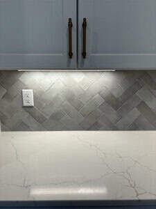 A kitchen counter features a white marble surface with subtle gray veining. The backsplash is composed of gray herringbone tiles. Above, white cabinets with gold handles are partially visible, and a power outlet is mounted on the wall.