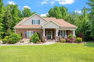 The charming house at 2053 Montgomery Drive in Franklinton features a stone facade, gable roof, and a front porch adorned with rocking chairs. It's surrounded by lush greenery and a well-maintained lawn, while an American flag hangs near the entrance on this bright, sunny day with blue sky and clouds.