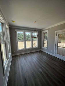 A bright, empty room with large windows allowing natural light to fill the space. The room features grey walls, dark wood flooring, a brass chandelier, and a glass door leading to a backyard or patio area.
