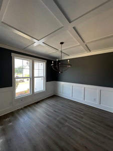 A bright dining room with dark gray walls, white wainscoting, and a coffered ceiling. A modern chandelier with six bulbs hangs from the ceiling. Large window allows natural light to fill the space. Wood flooring enhances the room's warmth.