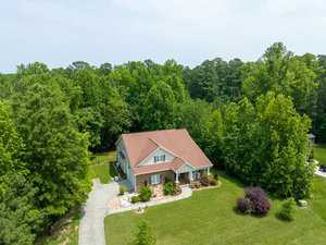 Aerial view of a suburban house with a red roof surrounded by lush green trees and a well-maintained lawn. A driveway leads to the front of the house, and there are garden beds along the path. The area is peaceful and verdant.