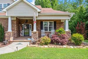 A cozy front porch with brick and stone detailing, featuring two white rocking chairs, a small table, and a potted plant. A lush green lawn and landscaped shrubs surround the area. An American flag hangs near the entrance.