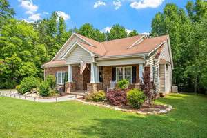 A charming one-story house with a mix of brick and siding exterior, adorned with an American flag. It features a neatly landscaped lawn, surrounded by lush green trees and a clear blue sky above.