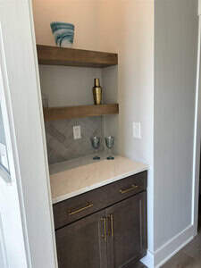 A small home bar area with a white marble countertop and a herringbone-patterned gray tile backsplash. Wooden shelves hold a decorative vase, brass shaker, and glasses. Below, a cabinet with brass handles offers additional storage.