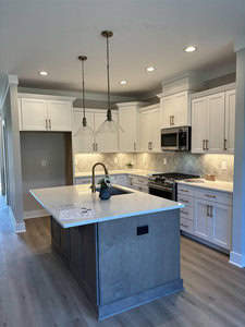 A modern kitchen with white cabinets, a gray island, wood flooring, and stainless steel appliances. Pendant lights hang over the island, and a herringbone tile backsplash accents the sleek, bright design.