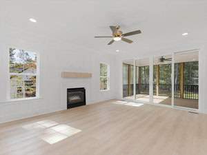 A bright, spacious living room with light wood flooring, a ceiling fan, a fireplace with a wooden mantel, and large sliding glass doors leading to a screened porch. Sunlight streams in through the windows and doors, illuminating the space.