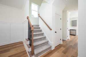 A bright, modern interior featuring a staircase with beige carpet and wooden handrails. The walls are light-colored, and the wood flooring extends into an adjacent room. A small window allows natural light to illuminate the entryway.