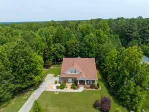 Aerial view of a single-story brick house with a brown roof, surrounded by lush green trees and a well-maintained lawn. A driveway leads to the house, and there are landscaped shrubs and plants in the front yard.