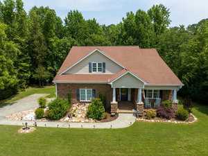 A charming one-story house with a brown roof and stone facade surrounded by lush greenery. The front yard features a neatly maintained lawn, decorative rocks, and shrubs. A concrete driveway leads to the entrance with chairs on the porch.