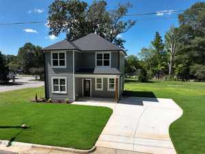 A two-story gray house with a dark roof is surrounded by a neatly cut lawn and positioned on a corner lot. A wide concrete driveway leads to the entrance, and tall trees fill the background under a clear blue sky.