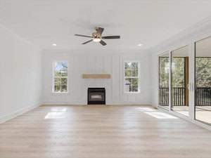 Spacious living room with light wood flooring, a ceiling fan, and a fireplace centered between two tall windows. Large glass sliding doors open to a covered balcony with views of greenery outside. Walls are painted white.