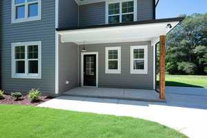A modern gray house with a small porch and white trim. The entrance features a black door with glass panels. The surrounding area includes a well-maintained green lawn, a concrete walkway, and a backdrop of lush trees.