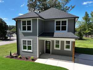 A modern, two-story house located at 608 Coggeshall Street in Oxford, with gray siding and a dark roof. It's surrounded by green grass and small shrubs. The home features large windows and a covered front porch with a glass door, set against trees and a clear blue sky.