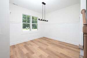 A bright, empty room with white walls and a light wood floor. A large window with a view of greenery is on the left wall. A black chandelier with candle-shaped bulbs hangs from the ceiling. Wainscoting decorates the lower walls.