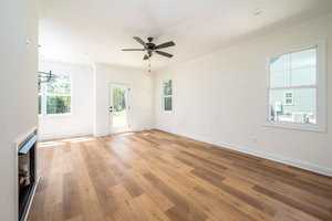 A bright, empty living room with light hardwood floors and pale walls. There's a ceiling fan and several windows. A door with a window leads outside, allowing natural light to fill the space.