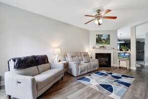 A cozy living room with a light gray sofa and recliners, a blue geometric rug, and a ceiling fan. A fireplace with a painting and aquarium sits in the corner. Soft lighting from a lamp and light walls add warmth to the space.