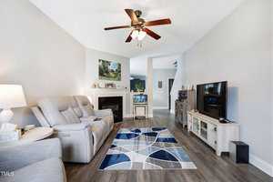 A modern living room with light gray walls and wooden flooring. It features a reclining sofa, a patterned blue and gray rug, a ceiling fan, a TV on a stand, and a fireplace with framed art above. Decorative items are on the shelves and mantel.