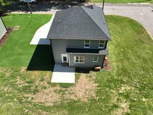 Aerial view of a two-story house with a dark roof and gray siding, surrounded by a mowed grassy yard. A driveway leads to the house, and a sidewalk outlines the property. The scene is set on a sunny day.