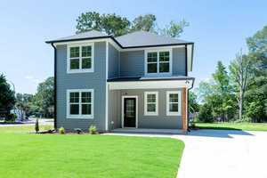 A modern two-story gray house with white trim and large windows. It features a covered porch, a driveway, and is surrounded by a well-maintained lawn and trees against a clear blue sky.