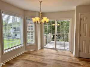 A bright dining area with hardwood floors, large windows, and a glass sliding door leading to a wooden deck. A chandelier hangs from the ceiling, and the windows offer a view of a lush green backyard.