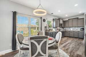 A modern kitchen features gray cabinets, a patterned tile backsplash, and a round dining table with four chairs centered on a rug. Large windows with gray curtains bring in natural light, illuminating the wood flooring.