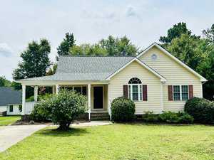 A charming yellow single-story house with white trim stands on 2752 Dorothy Court in Creedmoor. It features a gabled roof, front porch, and red shutters. Surrounded by a green lawn and shrubs, the home is set against a backdrop of trees under a partly cloudy sky.