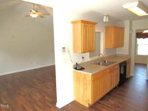 A kitchen with wooden cabinets and a double sink is shown. The countertop extends into a bar area. The room opens into a spacious living area with hardwood floors and a ceiling fan. Natural light filters through a window in the background.