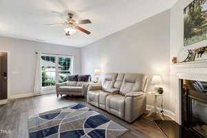 A modern living room with a gray reclining sofa and matching loveseat, a geometric patterned rug, and a ceiling fan. A fireplace is on the right, decorated with a clock and lamp. Large window with sheer curtains offers natural light and a view outside.