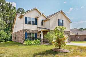 A two-story house with beige siding and brick accents, featuring blue shutters. It has a small, landscaped front yard with a tree and shrubs, and a paved driveway leading to a garage on the right. The background includes trees and a clear sky.