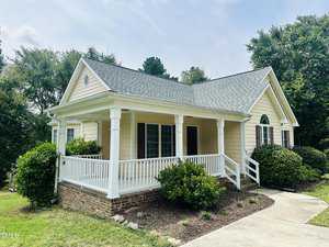 A small, single-story yellow house with a gray shingled roof and a covered porch. Surrounded by green shrubs and trees, it has a concrete walkway leading to the porch entrance. The sky is partly cloudy.