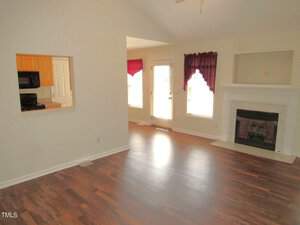 A bright living room with hardwood floors, a white fireplace, and two windows with red curtains. A door to the outside is between the windows. An open doorway leads to a kitchen with visible wooden cabinets.