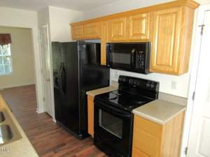 A kitchen featuring wooden cabinets, a black refrigerator, a black oven with an overhead microwave, and a double-basin stainless steel sink. The floor is wooden, and natural light comes in from a window in the background.