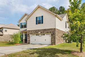 Two-story house with a mix of brick and siding exterior. The front features a double garage door and a small window above. A driveway leads to the garage, and a neatly maintained lawn surrounds the house, with a few trees nearby. Clouds fill the sky.