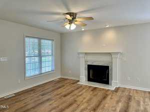 A bright living room with a ceiling fan, a large window with blinds, and a fireplace with a white mantel. The floor is made of light wood, and the walls are painted in a light, neutral color.