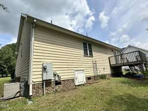 Side view of a single-story house with light-colored siding and a small deck. The house features a window, electrical meter, and an air conditioning unit. The lawn is neatly trimmed and a cloudy sky is visible overhead.
