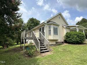 A light yellow house with white trim sits on a grassy lawn. It features a raised wooden porch with stairs leading up to it. There are bushes near the porch and trees in the background under a partly cloudy sky.