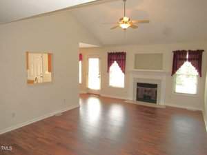 A spacious living room with vaulted ceilings, featuring hardwood floors, a ceiling fan, and a fireplace with a white mantel. Two windows with red curtains flank the fireplace. A door leads to an outside area, and there's an open pass-through to the kitchen.