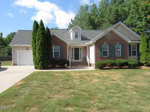 Located at 3639 Ashton Glen Lane, this charming Oxford-style abode is a brick and siding, single-story house with a gabled roof, flanked by tall evergreens and trimmed hedges. It features a covered entryway with steps, a front lawn, and an attached garage on the left. Blue shutters adorn the windows.