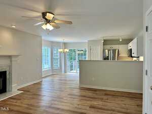 A spacious, open-concept living room with light wood flooring, a ceiling fan, and a fireplace on the left. It leads to a kitchen with white cabinets and stainless steel appliances. Large windows and glass doors provide ample natural light.